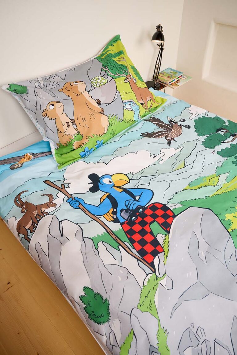 Die Kinderbettwäsche von Bettwäsche Globi Nationalpark zeigt ein Cartoon-Motiv mit einem blauen Vogel, der auf Felsen klettert, und zwei lächelnde braune Tiere im Gras sowie eine Lampe und Bücher auf dem Nachttisch.