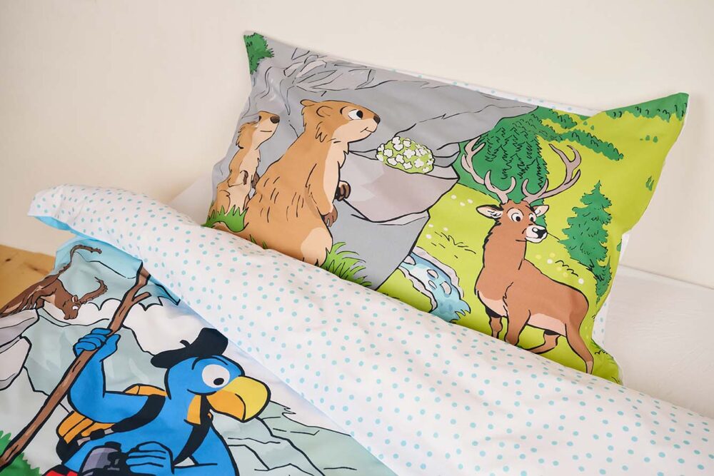 Das Bettwäsche Globi Nationalpark Set besteht aus einem Kissenbezug mit Murmeltieren und einem Reh in einem grünen Wald und einer Bettdecke mit Globi, dem blauen Vogel mit Hut und Rucksack, beide auf weißem Stoff mit blauen Punkten.