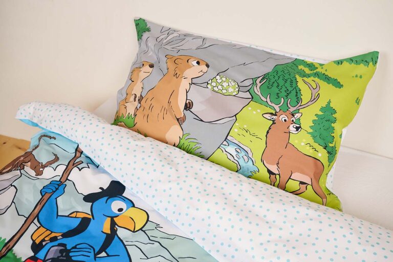 Das Bettwäsche Globi Nationalpark Set besteht aus einem Kissenbezug mit Murmeltieren und einem Reh in einem grünen Wald und einer Bettdecke mit Globi, dem blauen Vogel mit Hut und Rucksack, beide auf weißem Stoff mit blauen Punkten.