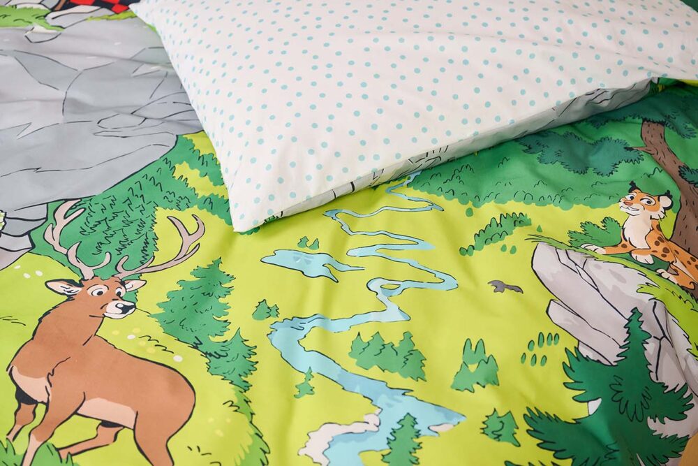 Die Bettwäsche Globi Nationalpark zeigt einen Cartoon-Hirsch und einen Luchs in einem lebendigen Wald an einem sich schlängelnden Fluss. Bäume und ein weißes Kissen mit blauen Tupfen vervollständigen das farbenfrohe Design.