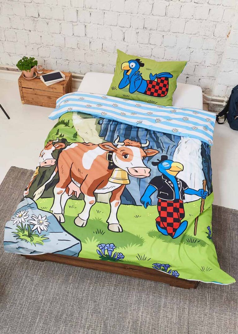 Das Bettwäsche Globi Alp Einzelbettset zeigt eine Kuh und einen Vogel in einer ländlichen Szene auf der Bettdecke, während das Kissen den Vogel mit einem Werkzeug vor einer rustikalen weißen Backsteinwand zeigt, mit einer kleinen Holzkiste neben dem Bett.