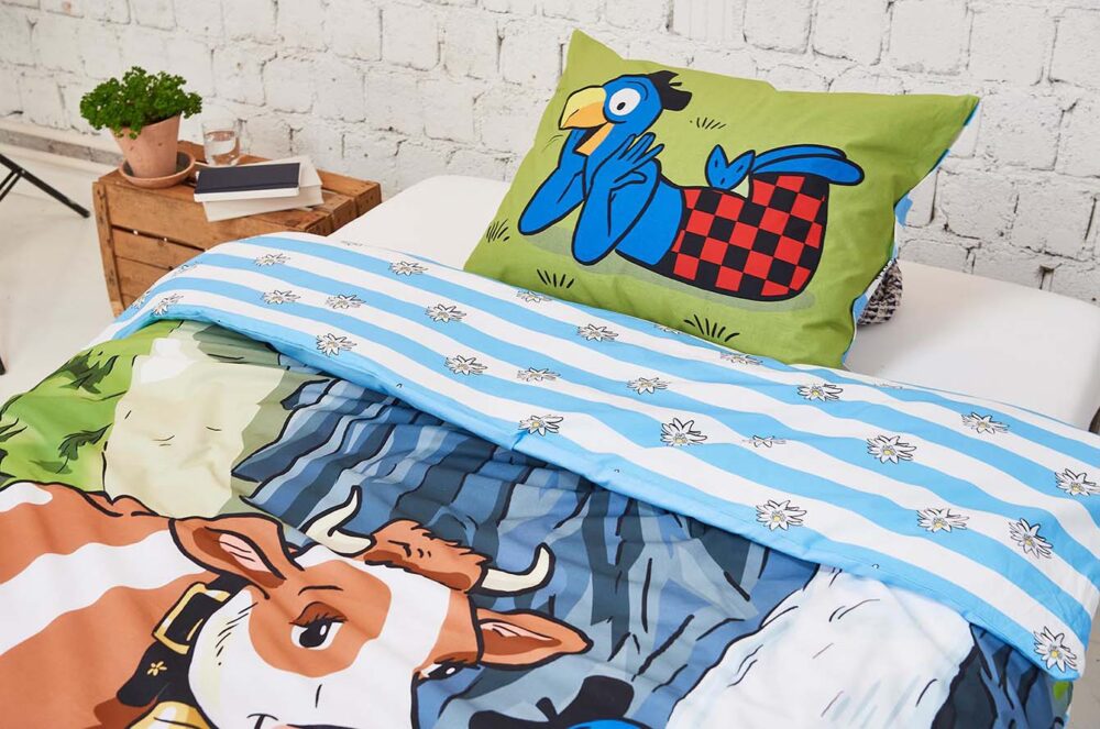 Ein Bett mit Bettwäsche Globi Alp - eine blau gestreifte Bettdecke mit einer Cartoon-Kuh und ein grünes Kissen mit einem blauen Cartoon-Vogel - steht neben einer weißen Ziegelwand und einer Holzkiste mit einer Topfpflanze.