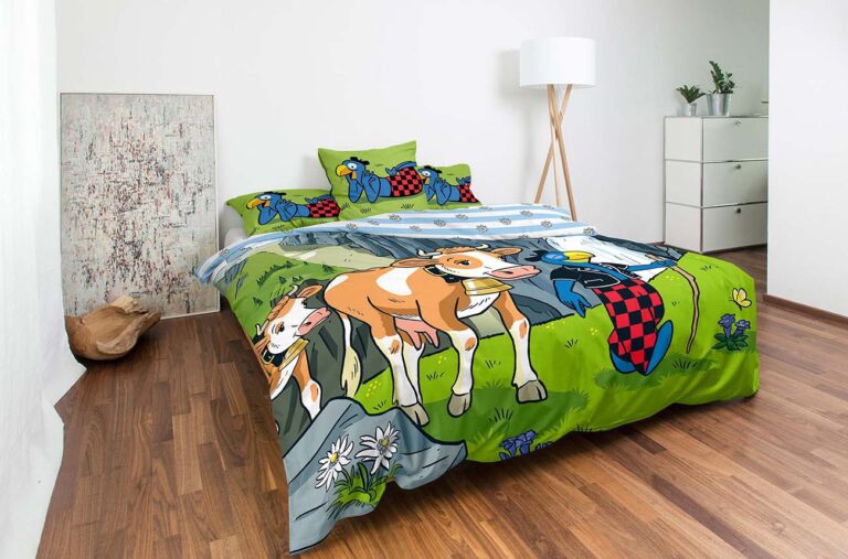 In einem modernen Schlafzimmer mit Holzböden, Topfpflanzen und einer weißen Lampe steht das Bettwäsche Globi Alp Set, das bunte Cartoon-Kühe und Bauernhofmotive auf einem ordentlich gemachten Bett zeigt.