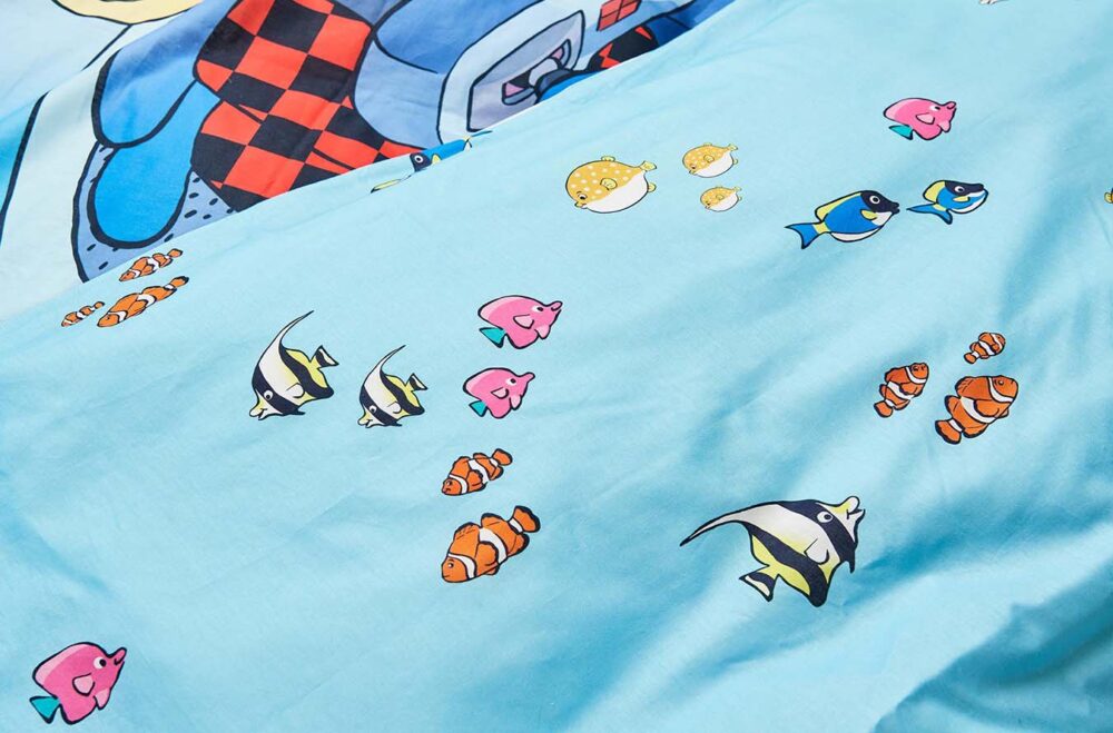 Eine Nahaufnahme des hellblauen Bettwäsche Globi Ozean-Stoffes zeigt bunte Cartoon-Fische, darunter Clownfische und Kaiserfische, mit einer größeren Cartoon-Figur und einem Schachbrettmuster im Hintergrund.