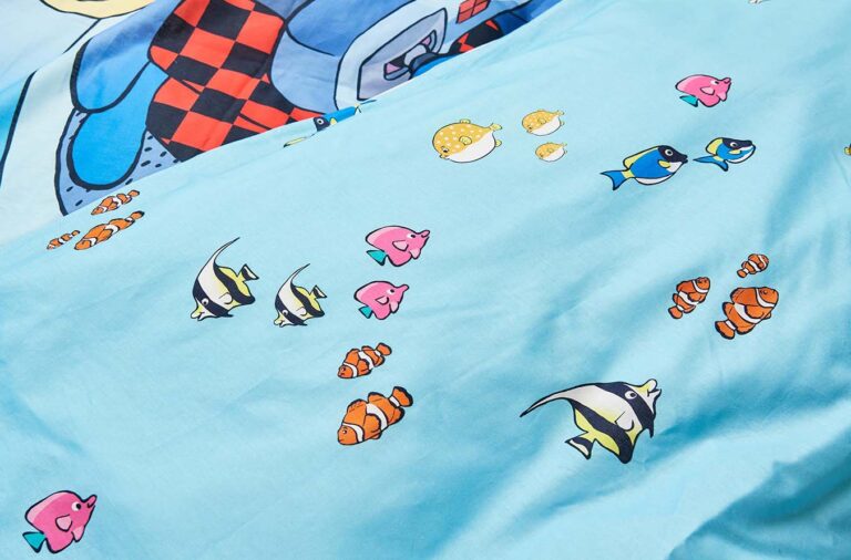 Eine Nahaufnahme des hellblauen Bettwäsche Globi Ozean-Stoffes zeigt bunte Cartoon-Fische, darunter Clownfische und Kaiserfische, mit einer größeren Cartoon-Figur und einem Schachbrettmuster im Hintergrund.