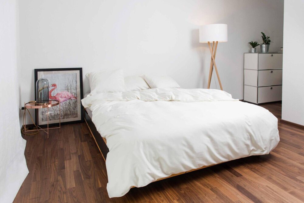 Ein minimalistisches Schlafzimmer mit einem weißen Bett mit Bettwäsche Nobilium Oni, einem Nachttisch, einer Stehlampe, rosa Flamingo-Kunst auf dem Boden und einer weißen Kommode mit grünen Pflanzen auf einem Holzfußboden.