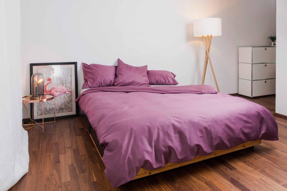Ein modernes Schlafzimmer mit Holzboden, einem Bett mit lila Bettwäsche Nobilium Oni, einer Stehlampe, einem runden Beistelltisch mit Flamingo-Kunstwerken und einem weißen Schubladenschrank vor sauberen weißen Wänden.