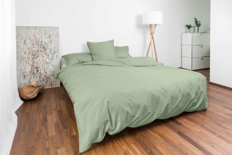 Ein modernes Schlafzimmer mit einem ordentlich gemachten Bett mit Bettwäsche Nobilium Oni in Salbeigrün, Holzböden, einer weißen Kommode mit Pflanzen, einer Stehlampe und abstrakter Kunst an der Wand.