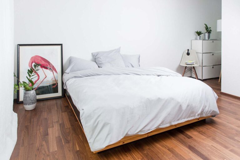 Minimalistisches Schlafzimmer mit einem niedrigen Holzbett, Bettwäsche Nobilium Oni in Hellgrau, einem gerahmten Flamingodruck auf dem Boden, Topfpflanzen, einer weißen Kommode und einem Stuhl mit Büchern auf Hartholzboden.