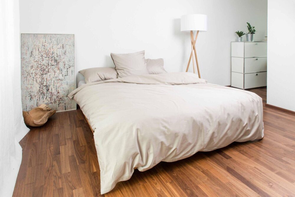 Ein minimalistisches Schlafzimmer mit einem sauber gemachten Bett in Bettwäsche Nobilium Oni, einer Stehlampe, einer Kommode mit Topfpflanzen, einem an die Wand gelehnten Strukturgemälde und einem Holzboden.