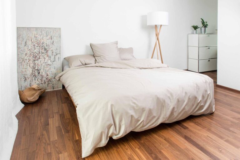 Ein minimalistisches Schlafzimmer mit einem sauber gemachten Bett in Bettwäsche Nobilium Oni, einer Stehlampe, einer Kommode mit Topfpflanzen, einem an die Wand gelehnten Strukturgemälde und einem Holzboden.