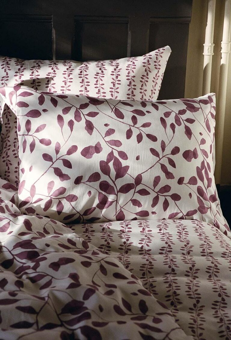 Ein Bett mit Bettwäsche Journey Living Pérouges - weiße Bettwäsche mit violetten Blättern und Zweigen - steht an einem Fenster mit beigen Vorhängen, das sanft vom natürlichen Licht erhellt wird.