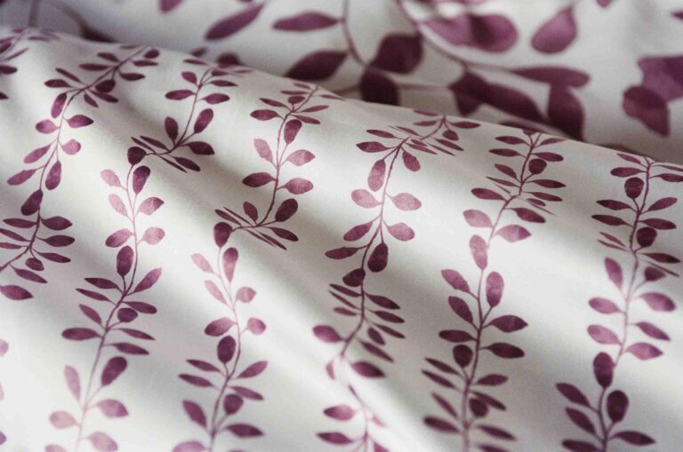 Eine Nahaufnahme von Bettwäsche Journey Living Pérouges zeigt den weißen Stoff mit einem sich wiederholenden kastanienbraunen Blatt- und Zweigmuster. Der sanft gefaltete Stoff erzeugt weiche Schatten und hebt das elegante botanische Muster hervor.