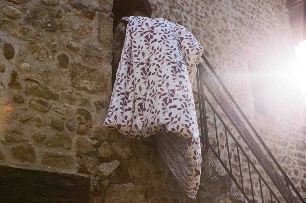 Die Bettwäsche Journey Living Pérouges mit braunem und violettem Blattmuster hängt über einer Steinwand und einer Metalltreppe im Sonnenlicht, während von rechts helles Licht einfällt.