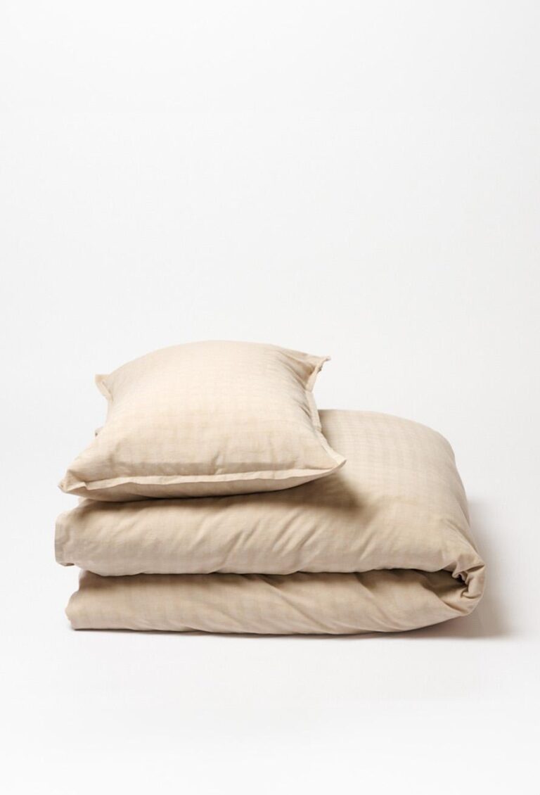 Ein beigefarbenes Kissen aus der Bettwäsche-Kollektion Journey Living Traboules ruht auf einer gefalteten Bettdecke, beide mit einem dezenten Karomuster vor einem schlichten weißen Hintergrund.
