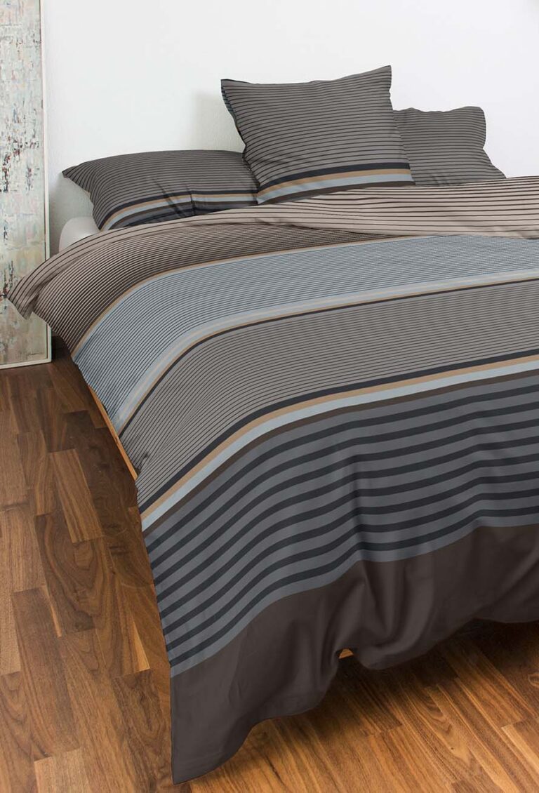 Ein ordentlich gemachtes Bett mit Bettwäsche Night Fashion Orsa in gestreiften Grau-, Braun- und Schwarztönen und passenden Kissen steht auf einem Holzboden neben einer hellen Wand.