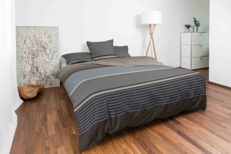 Ein modernes, minimalistisches Schlafzimmer mit Holzfußboden verfügt über ein ordentlich gemachtes Bett in grau gestreifter Bettwäsche Night Fashion Orsa, ergänzt durch eine Stehlampe, eine Kommode, eine Pflanze und ein abstraktes Gemälde, das auf dem Boden liegt.