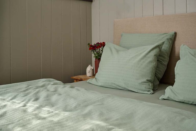 Ein gemütliches Bett mit Bettwäsche Journey Living Kemeri in Hellgrün und passenden Kissen steht neben einem hölzernen Nachttisch, auf dem eine Vase mit roten Blumen und Dekoration steht, vor hell getäfelten Wänden.