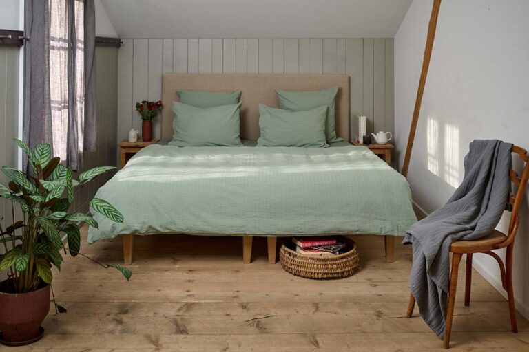 Das gemütliche Schlafzimmer verfügt über ein großes Bett mit mintgrüner Bettwäsche von Journey Living Kemeri, zwei hölzerne Nachttische, Pflanzen, einen in eine graue Decke gehüllten Stuhl und helle Holzböden, die in sanftes Tageslicht getaucht sind.
