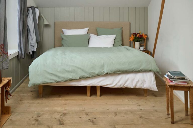 Ein gemütliches Schlafzimmer mit einem ordentlich gemachten Bett mit Bettwäsche Journey Living Kemeri in Grün und Weiß, zwei grünen Kissen, einem beigen Kopfteil und einem hölzernen Nachttisch mit Büchern, Blumen und einer Tasse auf dem hellen Holzboden.