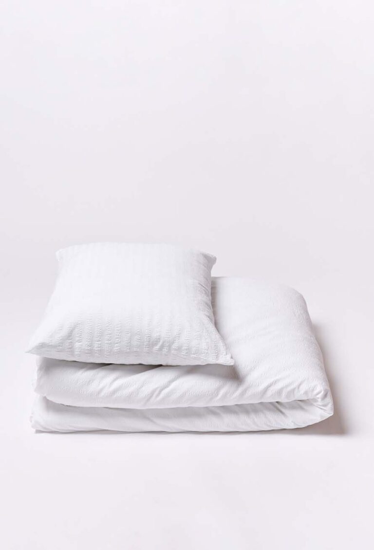 Ein weißes Kissen liegt auf einer ordentlich gefalteten weißen Bettdecke von Bettwäsche Journey Living Kemeri, beide vor einem schlichten weißen Hintergrund.