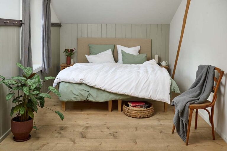 Ein gemütliches Schlafzimmer mit einem großen Bett mit Bettwäsche Journey Living Kemeri in Grün und Weiß, Büchern in einem Korb unter dem Bett, einer Topfpflanze, einem mit einer grauen Decke drapierten Holzstuhl und weichem, natürlichem Fensterlicht.