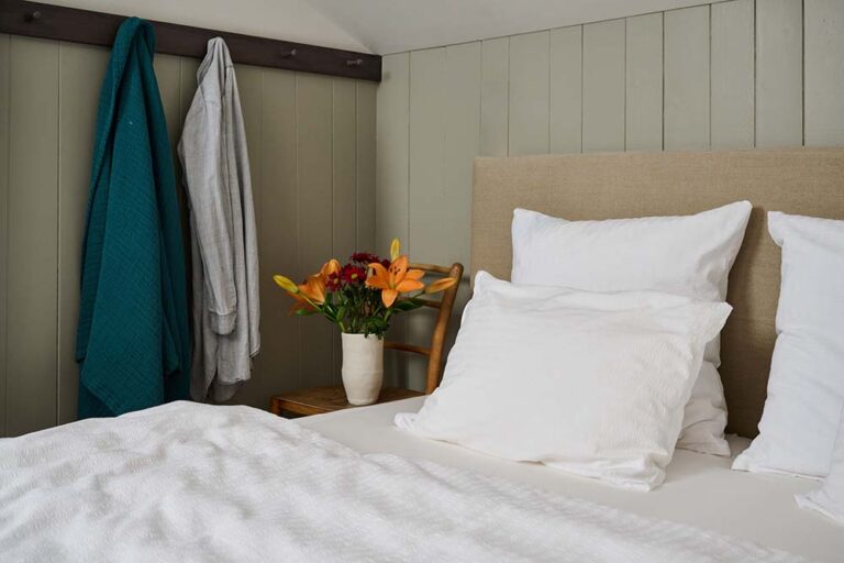 Das gemütliche Schlafzimmer verfügt über ein Bett mit weißer Bettwäsche von Journey Living Kemeri, flauschige Kissen, ein hellbraunes Kopfteil, einen Holzstuhl als Nachttisch mit frischen Blumen und zwei Bademäntel, die an einer hellgrün getäfelten Wand hängen.