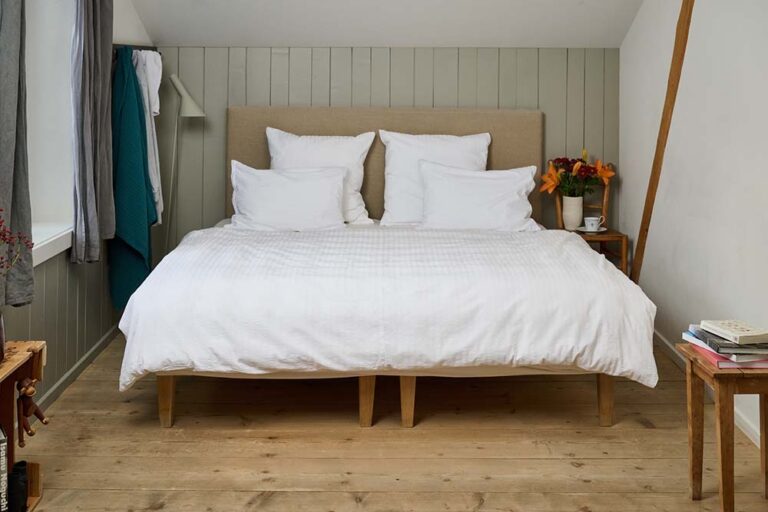Ein gemütliches Schlafzimmer mit einem ordentlich gemachten Bett in Bettwäsche Journey Living Kemeri, vier weißen Plüschkissen, Holzfußboden, Nachttischen mit Büchern, einer Blumenvase und einer Teetasse. Hell getäfelte Wände rahmen den ruhigen Raum ein.