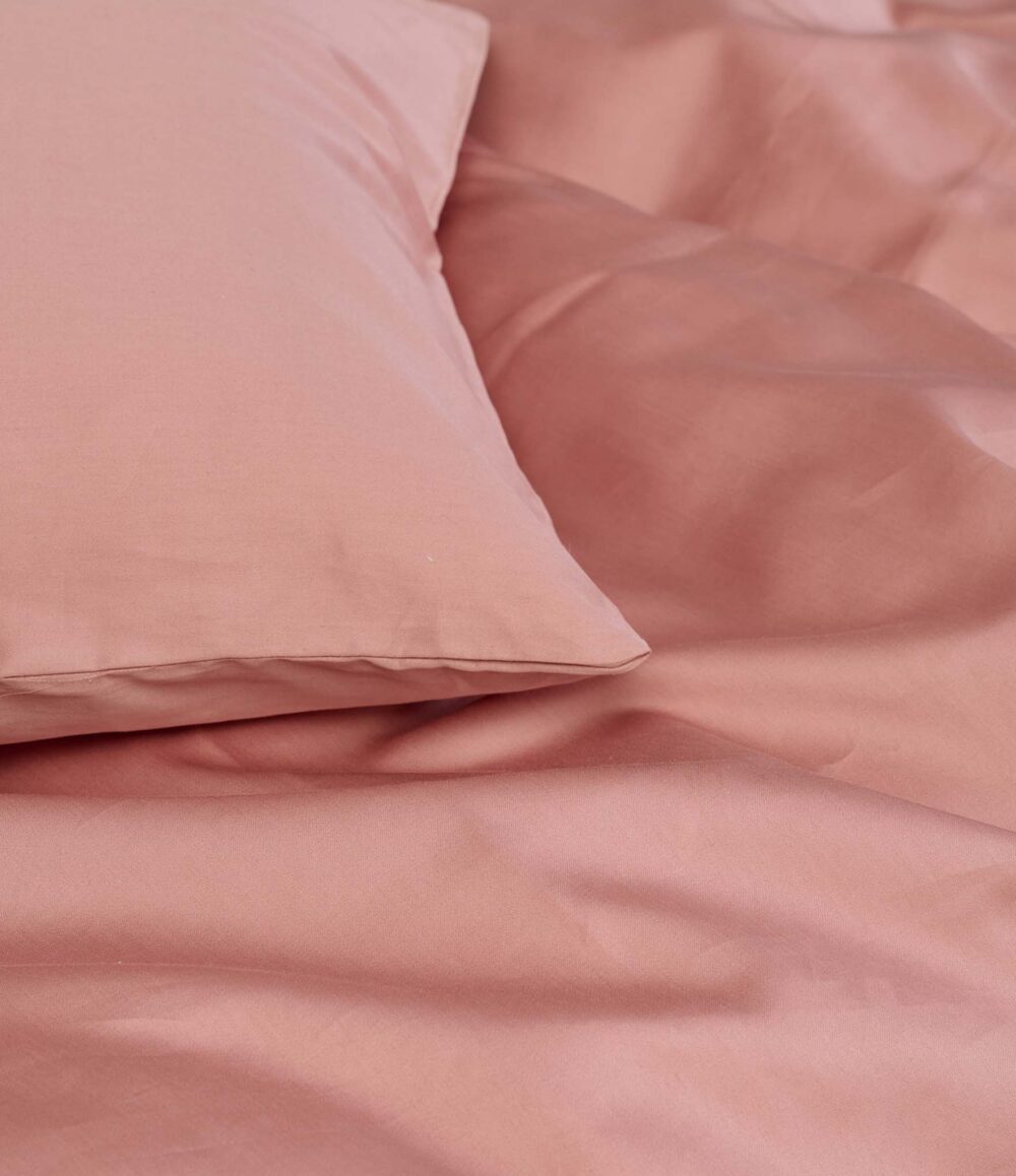 Die Nahaufnahme eines gemachten Bettes zeigt ein rosafarbenes Kissen und die Bettwäsche Braga Dusty Powder von Journey Living. Beide sind aus glattem Stoff mit weichen Falten gefertigt und schaffen eine ruhige und gemütliche Atmosphäre.
