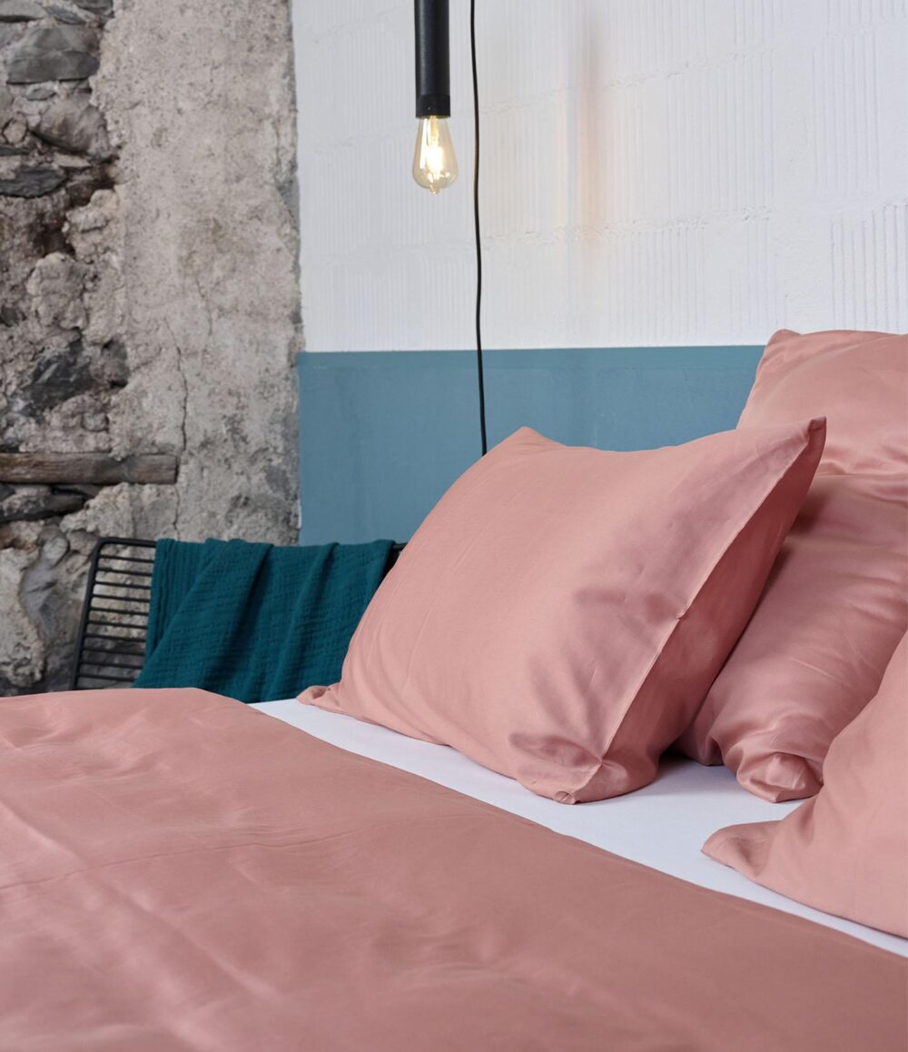 Bettwäsche Journey Living Braga Dusty Powder Laken und Kissen auf einem Bett an einer rustikalen Steinwand, mit einer dunkelgrünen Decke auf einem schwarzen Metallstuhl und einer freiliegenden Glühbirne darüber.