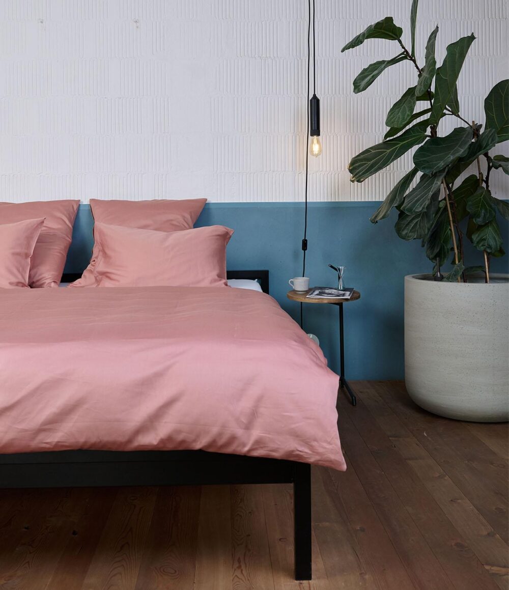 In einem modernen Schlafzimmer stehen ein Bett mit Bettwäsche Journey Living Braga Dusty Powder, ein kleiner runder Beistelltisch mit einer Tasse und Büchern, eine Hängelampe und eine große Topfpflanze vor blauen und weißen Wänden.