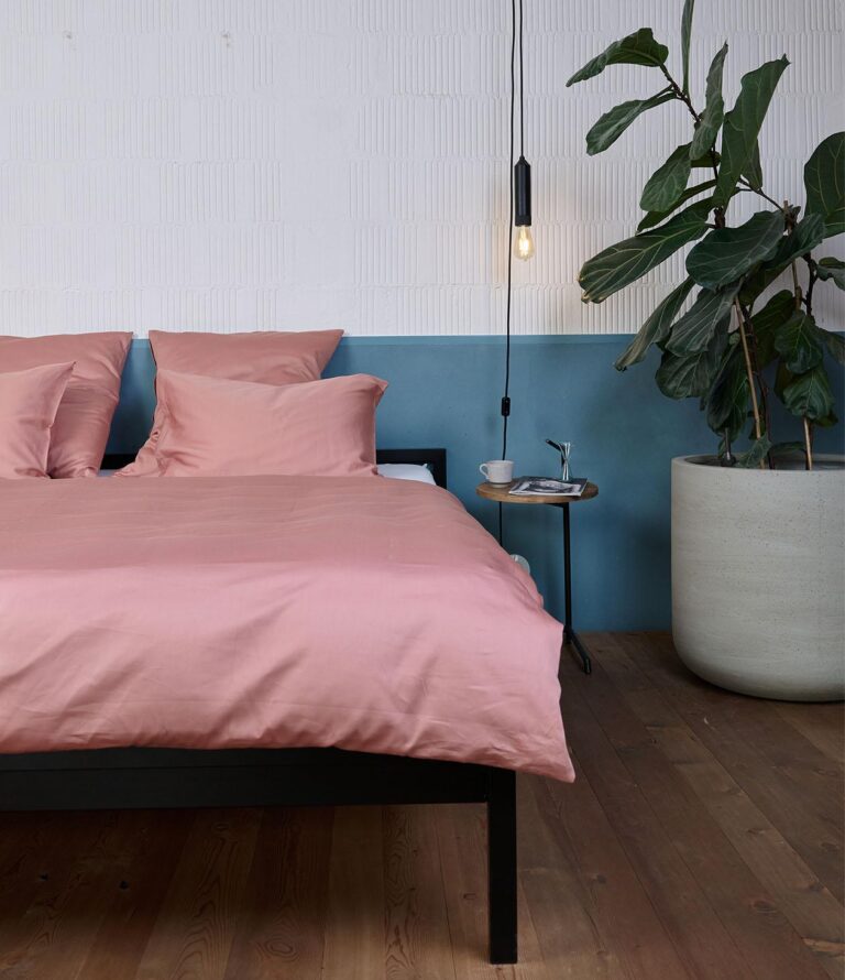 In einem modernen Schlafzimmer stehen ein Bett mit Bettwäsche Journey Living Braga Dusty Powder, ein kleiner runder Beistelltisch mit einer Tasse und Büchern, eine Hängelampe und eine große Topfpflanze vor blauen und weißen Wänden.