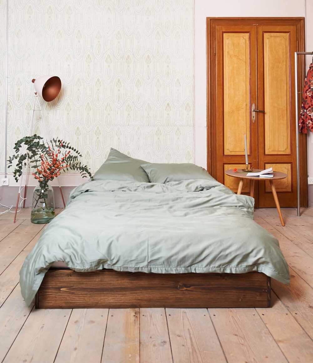 Ein gemütliches Schlafzimmer mit einem Bettgestell aus Holz und grüner Bettwäsche Journey Living Braga Eucalyptus und Kissen. Auf einem runden Beistelltisch aus Holz stehen Kerzen, eine moderne Lampe und eine Pflanze. Hölzerne Flügeltüren und gemusterte Tapeten vervollständigen den Look.