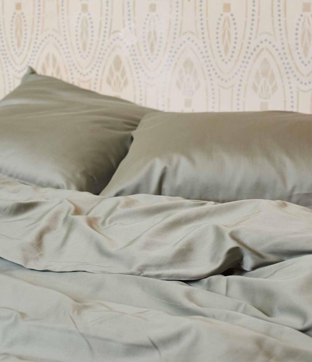 Zwei Kissen und eine Bettdecke mit der glatten, hellsalbeigrünen Bettwäsche Journey Living Braga Eucalyptus sind auf einem Bett vor cremefarbener Mustertapete angeordnet.