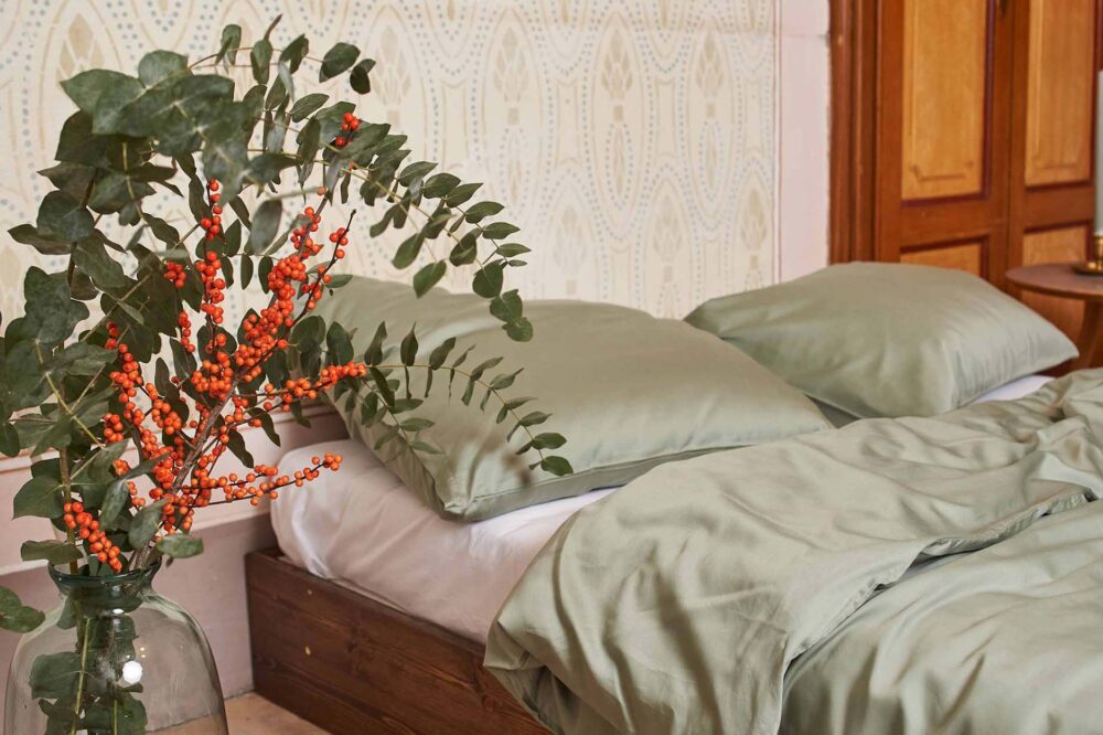 Ein gemütliches Bett mit salbeigrünem Bettwäsche Journey Living Braga Eukalyptus und Kissen steht vor einer gemusterten Wand. Daneben steht eine Glasvase mit grünen Eukalyptuszweigen und leuchtend orangefarbenen Beerenbüscheln.
