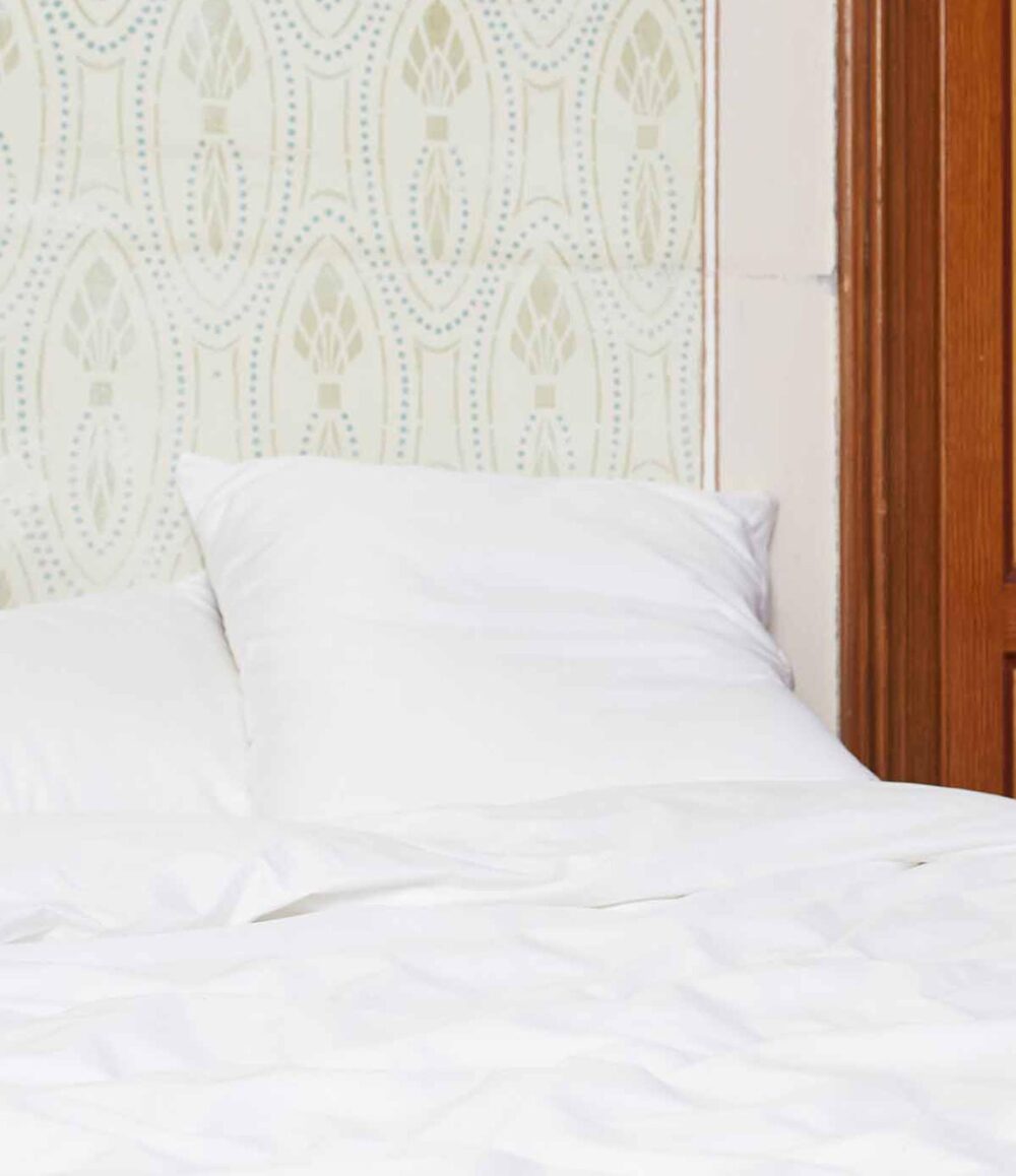 Ein ordentlich gemachtes Bett mit weißer Bettwäsche Journey Living Braga Snow und Kissen, bedeckt mit einer weißen Decke, vor einer gemusterten Tapete neben einer Holztür oder -platte.