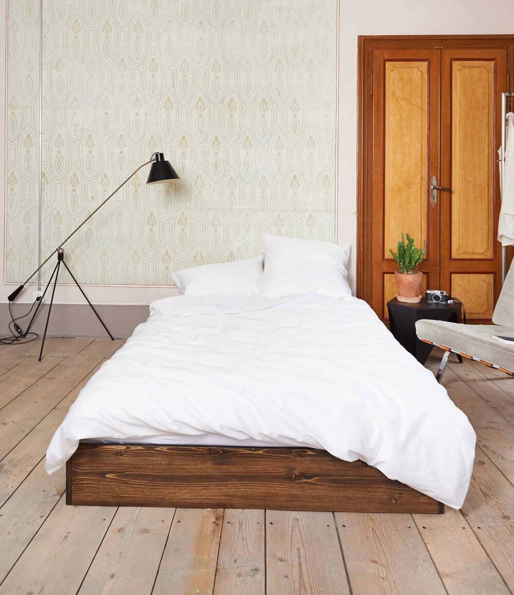 Ein einfaches Schlafzimmer mit einem Holzbettgestell, Bettwäsche Journey Living Braga Snow und zwei Kissen, einer schwarzen Stehlampe, einem kleinen Beistelltisch mit einer Pflanze, einem Stuhl, Flügeltüren aus Holz, einem hellen Holzboden und einer dezent gemusterten Tapete.