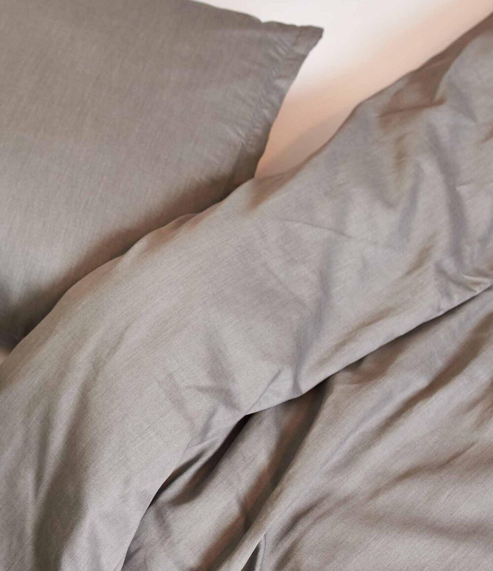 Nahaufnahme eines Bettes mit Bettwäsche Journey Living Braga Stone und passendem Kopfkissen. Hervorzuheben sind die hellgraue Farbe und die leicht faltige Stoffstruktur.