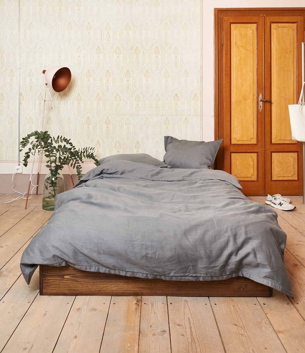 Ein ordentlich gemachtes Bett mit Bettwäsche Journey Living Braga Stone steht auf einem Holzboden, dahinter eine Blattpflanze, eine Stehlampe und eine gemusterte Tapete. Auf der rechten Seite befinden sich hölzerne Doppeltüren und weiße Turnschuhe.