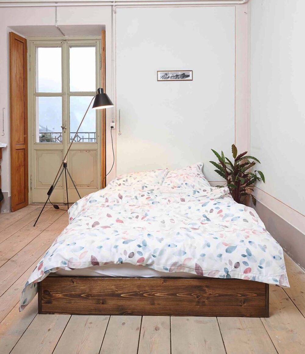Das gemütliche Schlafzimmer ist mit einem Holzbettgestell mit Bettwäsche Journey Living Greina in einem bunten Blattmuster, einer hohen Stehlampe, einer Topfpflanze und einem großen Fenster mit Holzläden ausgestattet, das natürliches Licht hereinlässt.