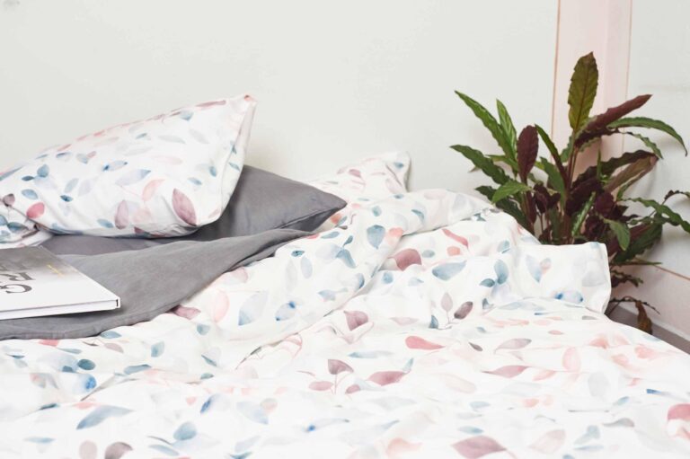 Ein Bett mit weißen Laken und Kissenbezügen von Bettwäsche Journey Living Greina mit pastellfarbenem Blattmuster, ein graues Kissen, ein aufgeschlagenes Buch und eine grüne Topfpflanze stehen daneben an einer weißen Wand.