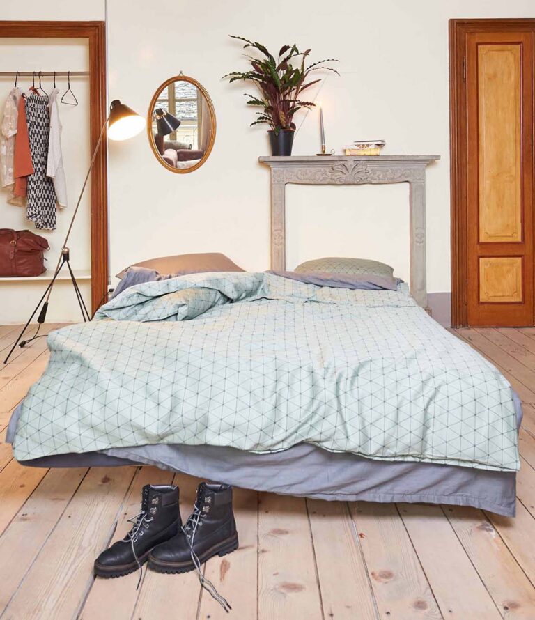 Ein ordentlich gemachtes Bett mit Bettwäsche Journey Living Peniche und einer geometrisch gemusterten Bettdecke steht in einem gemütlichen Schlafzimmer. Schwarze Stiefel stehen auf dem Holzboden, eine Lampe, hängende Kleidung, ein runder Spiegel und eine Topfpflanze ergänzen die Einrichtung.