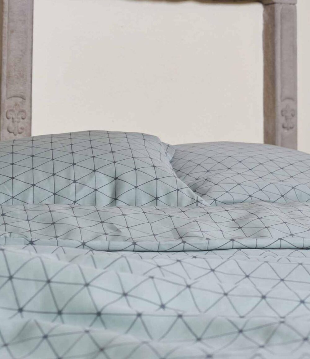 Die Nahaufnahme eines ordentlich gemachten Bettes zeigt die Bettwäsche Journey Living Peniche in Hellblau mit schwarzem geometrischem Gittermuster, gepaart mit einem Holzkopfteil im Hintergrund.