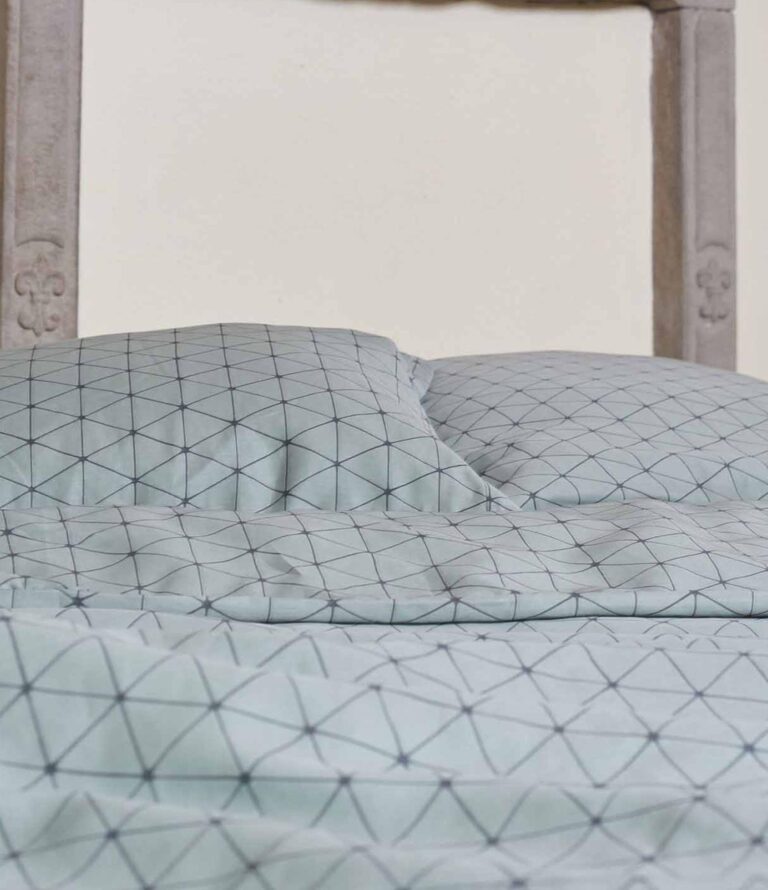 Die Nahaufnahme eines ordentlich gemachten Bettes zeigt die Bettwäsche Journey Living Peniche in Hellblau mit schwarzem geometrischem Gittermuster, gepaart mit einem Holzkopfteil im Hintergrund.