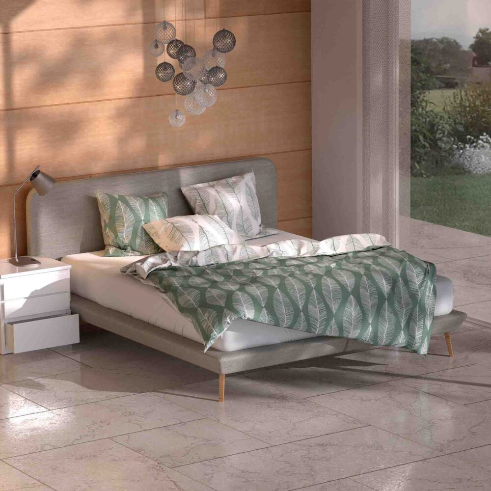 Ein modernes Schlafzimmer mit einem grauen Polsterbett, Bettwäsche Ruben Lotus Création mit grünem und weißem Blattdruck, zwei weißen Nachttischen, einer Schreibtischlampe, kreisförmiger Wandkunst und Sonnenlicht auf dem glänzenden Fliesenboden.