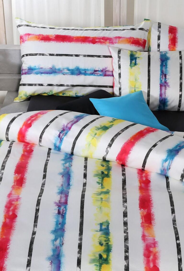 Ein Bett ist mit der Bettwäsche Tamara-R Selection Laurel bezogen, die aus weißer Bettwäsche mit schwarzen, roten, blauen, gelben und grünen Querstreifen im Aquarellstil besteht; ein hellblaues Akzentkissen hebt sich von den gemusterten Kissen ab.
