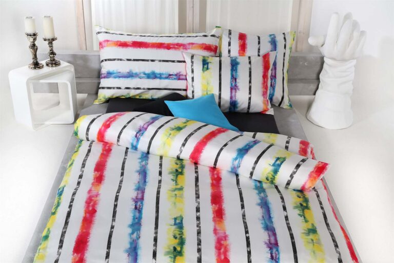 Ein Bett mit der farbenfrohen Bettwäsche Tamara-R Selection Laurel in Rot, Gelb, Blau und Schwarz, zwei Kissen, ein Beistelltisch mit zwei hohen Kerzen und eine weiße dekorative Handskulptur neben dem Bett.