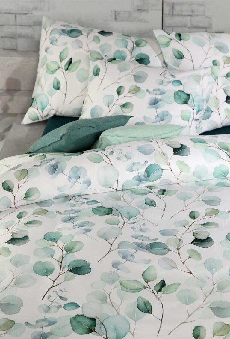 Ein Bett mit weißer Bettwäsche und Kissen zeigt das blattgrüne und blaue Aquarellmuster von Bettwäsche Tamara-R Selection Benita vor einer hellgrauen Backsteinwand.
