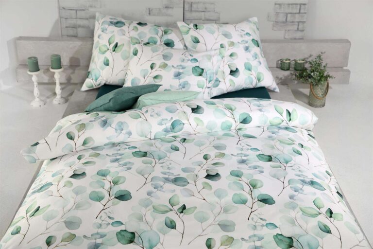 Das sauber gemachte Bett ist mit Bettwäsche Tamara-R Selection Benita und Kopfkissenbezügen mit grünem Blattmuster bezogen, ergänzt durch dekorative Kerzen und eine Topfpflanze auf dem Nachttisch.