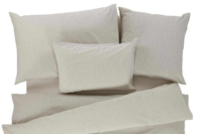 Ein ordentlich gemachtes Bett mit hellbeiger Bettwäsche Tamara-R BeCosy Lee, zwei großen Kissen, einem mittelgroßen Kissen und einer Bettdecke auf einer flachen Oberfläche.